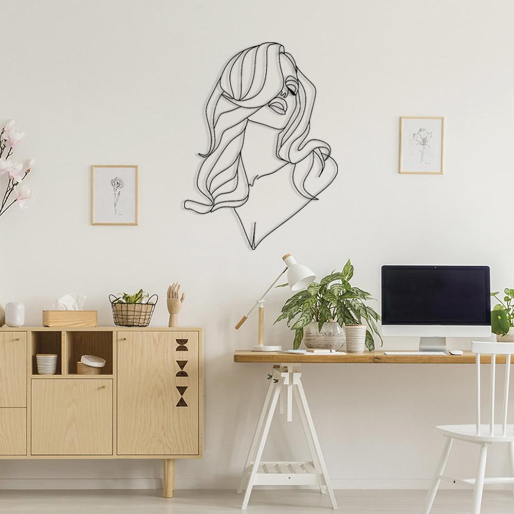 Sleek line art metal wall decor of a woman with flowing long hair mounted on a white wall in a minimalist study area. The room features a wooden desk with a white trestle frame, a computer setup, indoor plants, and a Scandinavian-style sideboard with decorative items and framed art.