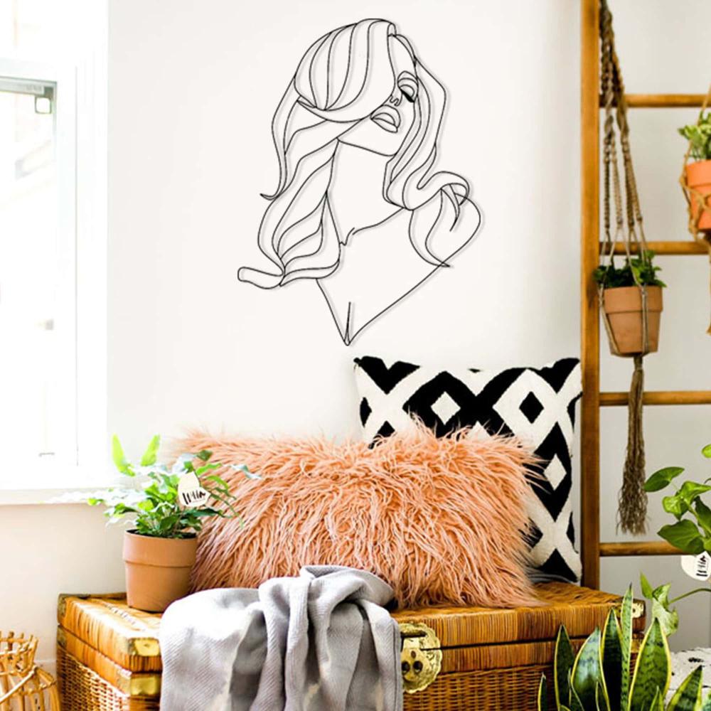 Stylish metal wall art featuring the silhouette of a woman with long flowing hair, mounted on a light-colored wall above a cozy seating area adorned with plants, pillows, and a throw blanket.