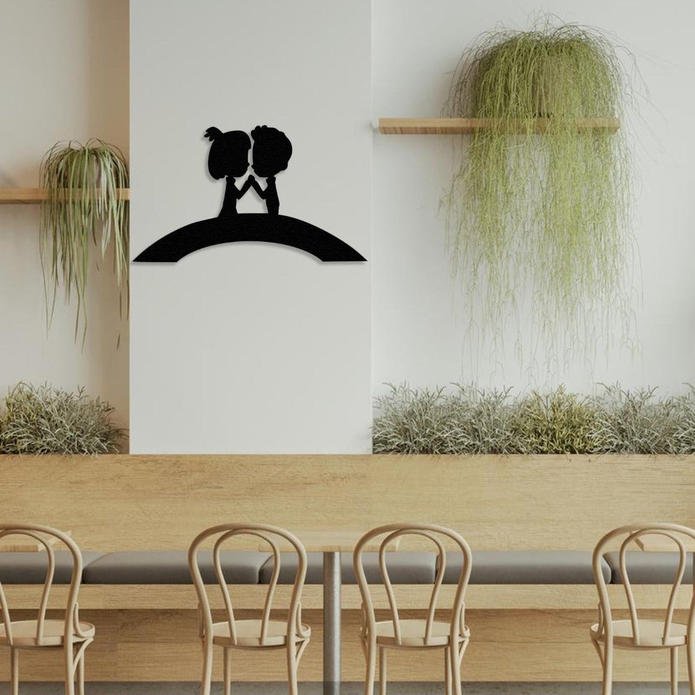 Silhouette-style metal wall art featuring a couple holding hands on a bridge, displayed in a minimalist café with wooden furniture and lush greenery.
