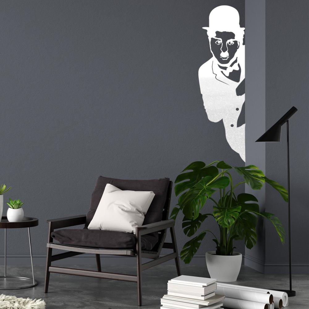 White metal Charlie Chaplin wall art featuring a silhouette of the iconic figure peeking from behind a corner, mounted on a dark gray wall above a modern armchair with a pillow, alongside a potted plant and a sleek lamp.