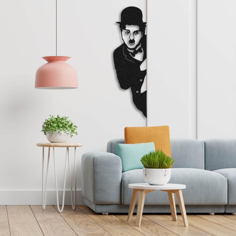 Black Charlie Chaplin metal wall art mounted on a white wall, positioned near a modern gray sofa with colorful cushions, a wooden side table with a plant, and a pink pendant lamp adding a vibrant touch to the decor.
