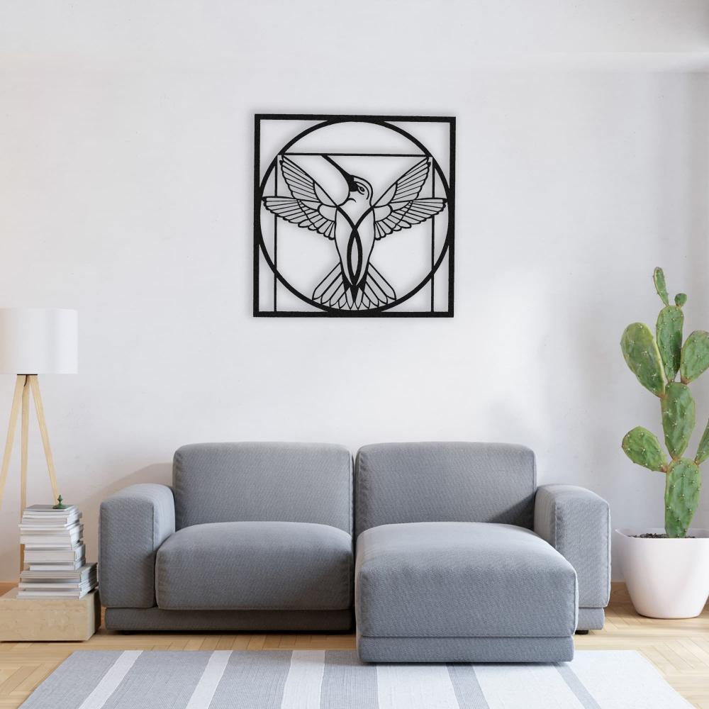 Black metal hummingbird wall art with a geometric circular design displayed above a modern gray sofa in a minimalist living room with a cactus plant and a tripod lamp.