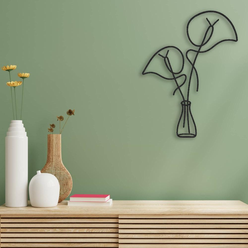 Black metal wall art featuring a minimalist design of leaves in a vase, mounted on a green wall above a wooden console table. The console is decorated with white ceramic vases, a wicker vase, and stacked books, adding a natural touch to the decor.