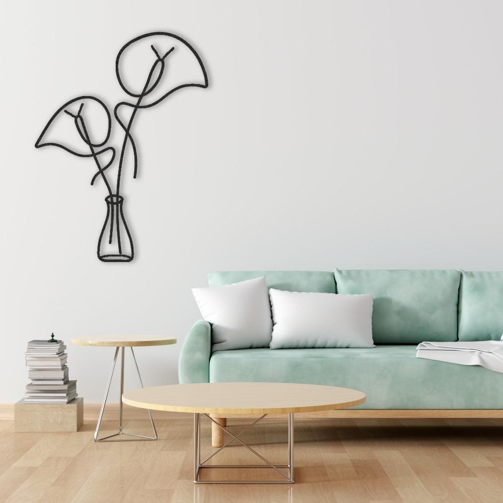 Black metal wall art depicting minimalist leaves in a vase, mounted on a light-colored wall. Below is a light green sofa with white pillows, accompanied by round wooden coffee and side tables, and a stack of books for a modern, cozy living room setup.