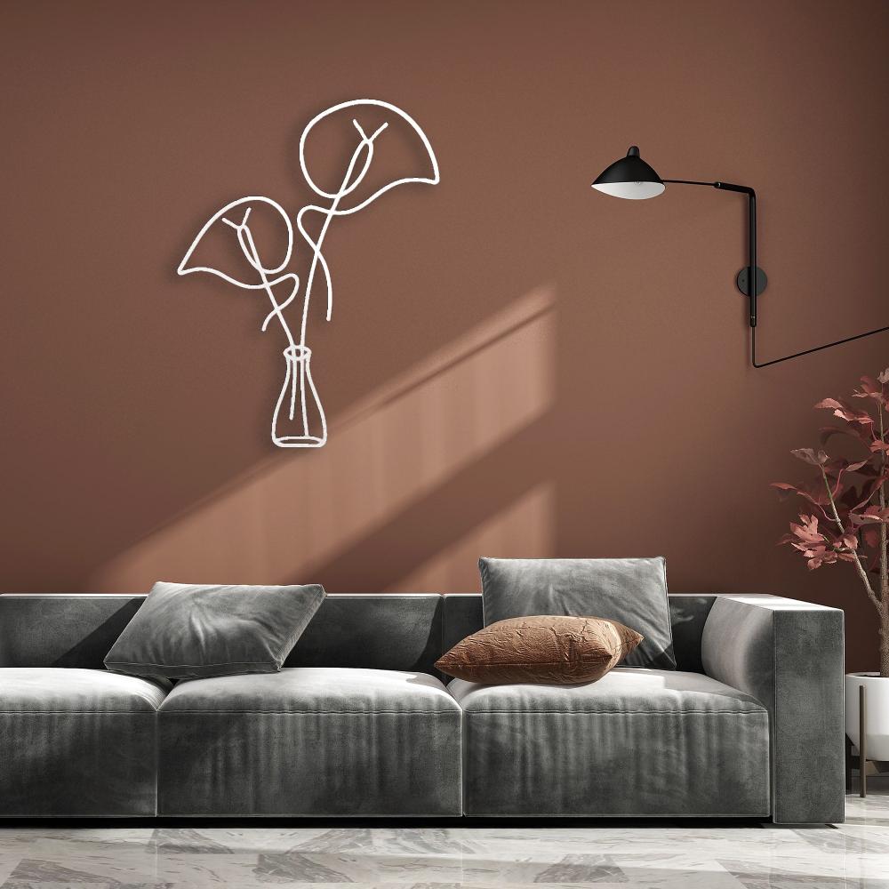 White minimalist metal wall art featuring abstract leaves in a vase, mounted on a warm brown wall. Positioned above a modern gray velvet sofa with a textured brown throw pillow, complemented by a sleek black wall lamp and a decorative potted plant in the corner.