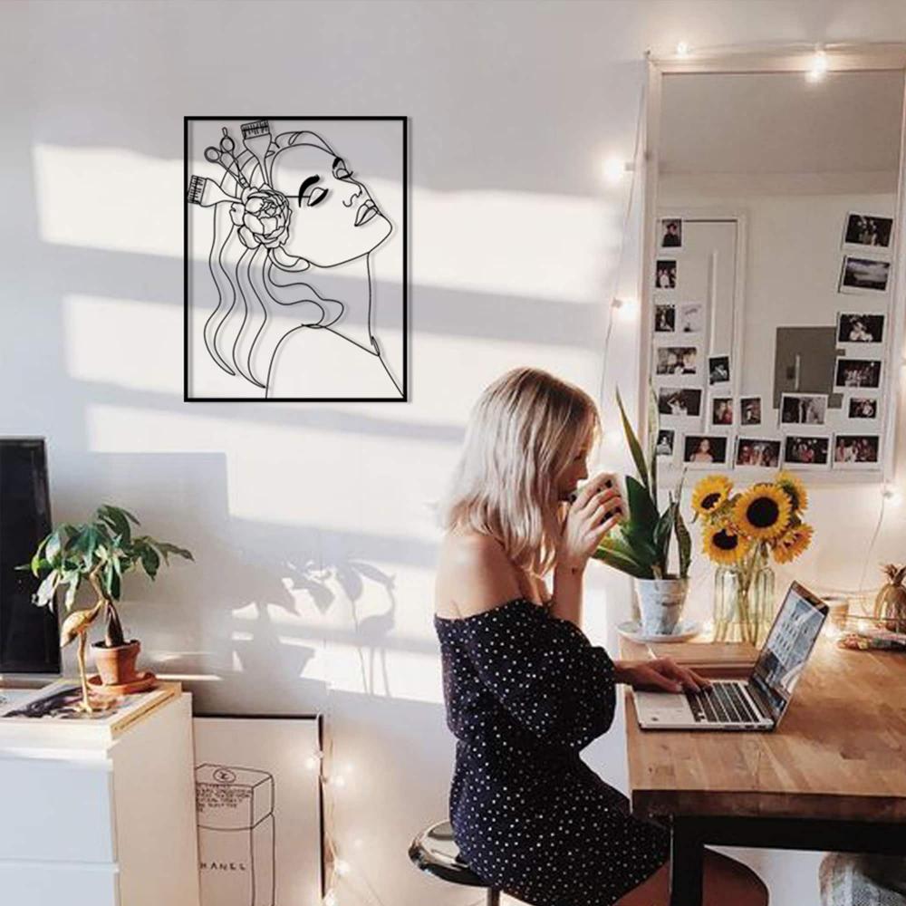 A modern workspace with black metal wall art of a woman’s profile with hairdressing tools and a flower, mounted on the wall behind a desk with a woman working on a laptop.