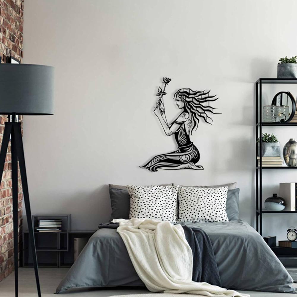 Modern bedroom featuring black metal wall art of a woman holding a rose, mounted above a gray bed with patterned pillows and a cozy white throw blanket. The room includes a tall black floor lamp, exposed brick wall, and sleek black shelving with decorative elements.