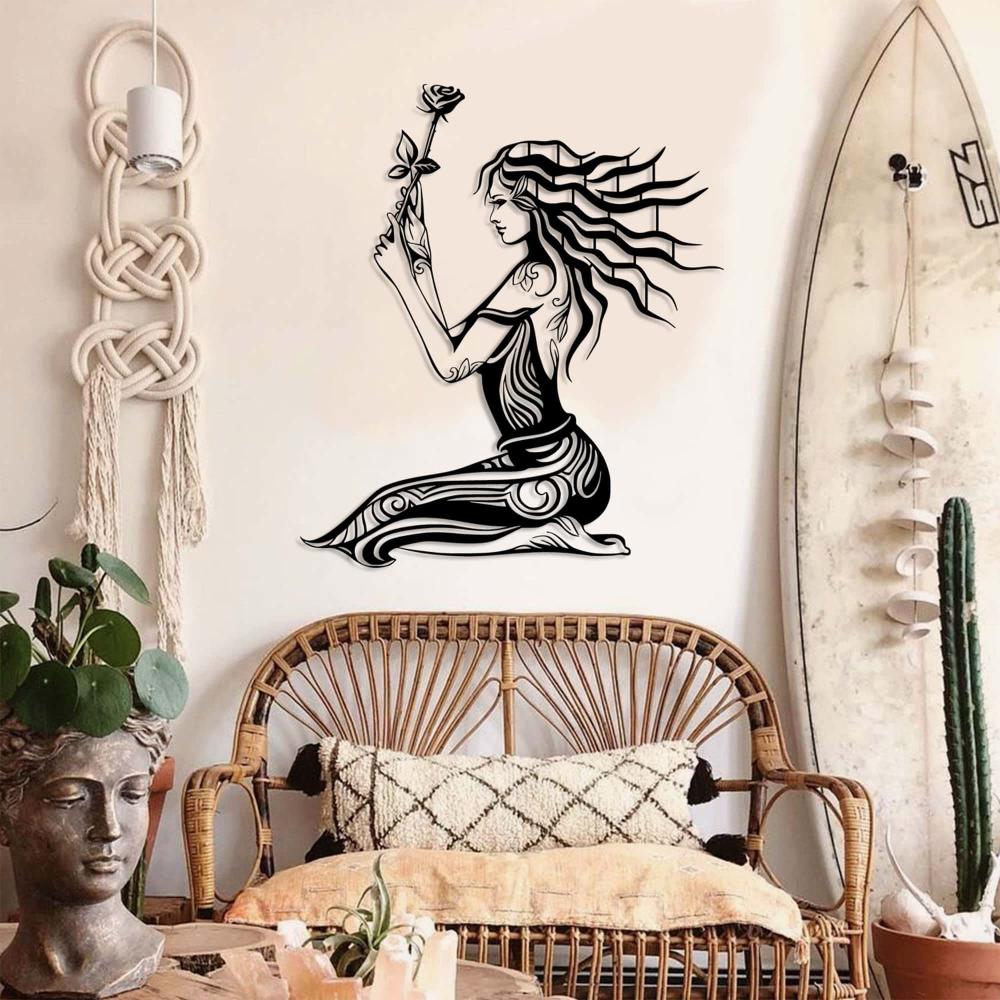 Elegant black metal wall art of a seated woman holding a rose, featuring intricate swirling patterns and flowing hair, displayed in a bohemian-style room. The decor includes a rattan bench with patterned cushions, a macrame wall hanging, a surfboard leaning against the wall, potted plants, and a decorative bust.
