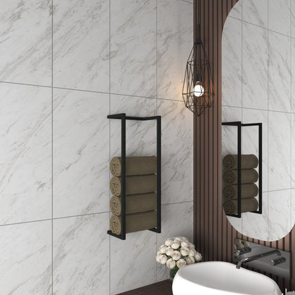 Black wall-mounted towel rack holding four rolled towels, installed in a modern bathroom with marble tiled walls, a stylish pendant light, and a round mirror next to a sink with white flowers below.