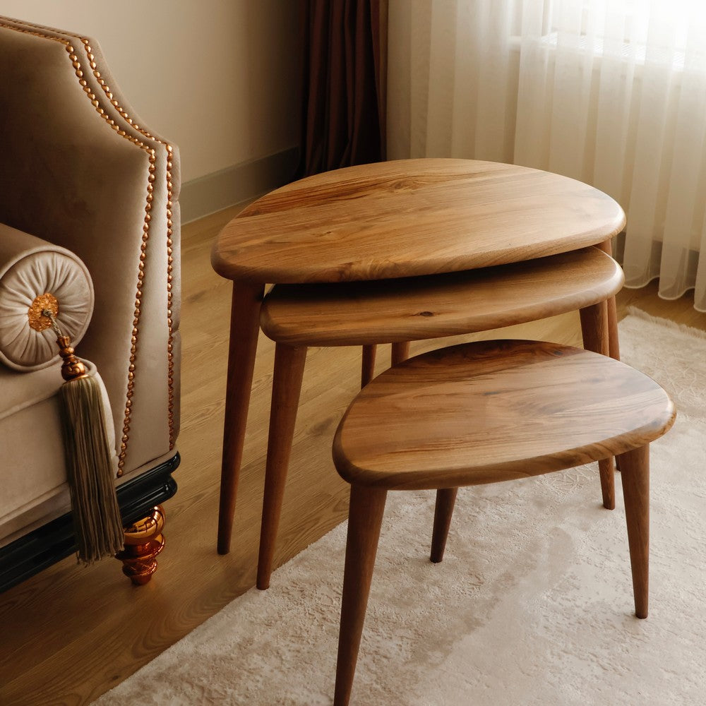 Elegant set of three oval wooden nesting tables with tapered legs, placed next to a luxurious beige armchair.