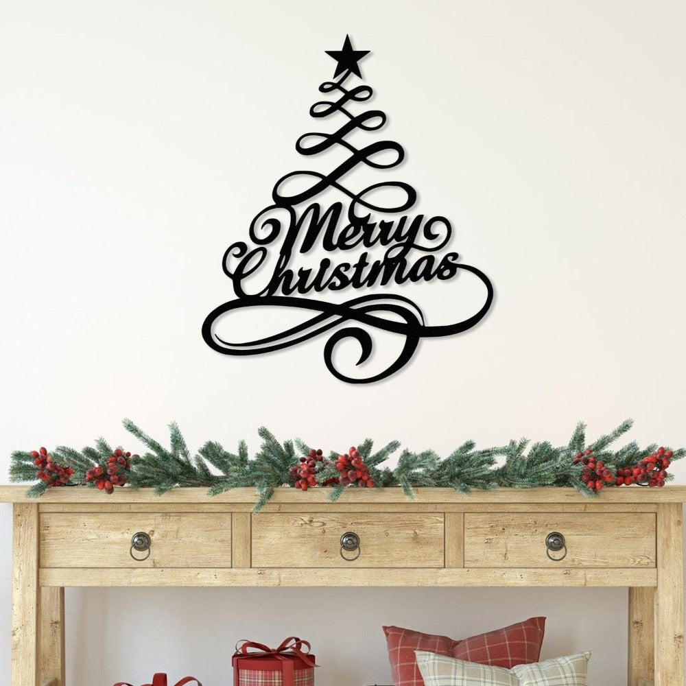 Black metal "Merry Christmas" wall art shaped like a Christmas tree with a star topper, elegantly displayed above a rustic wooden table decorated with festive greenery and red berries.