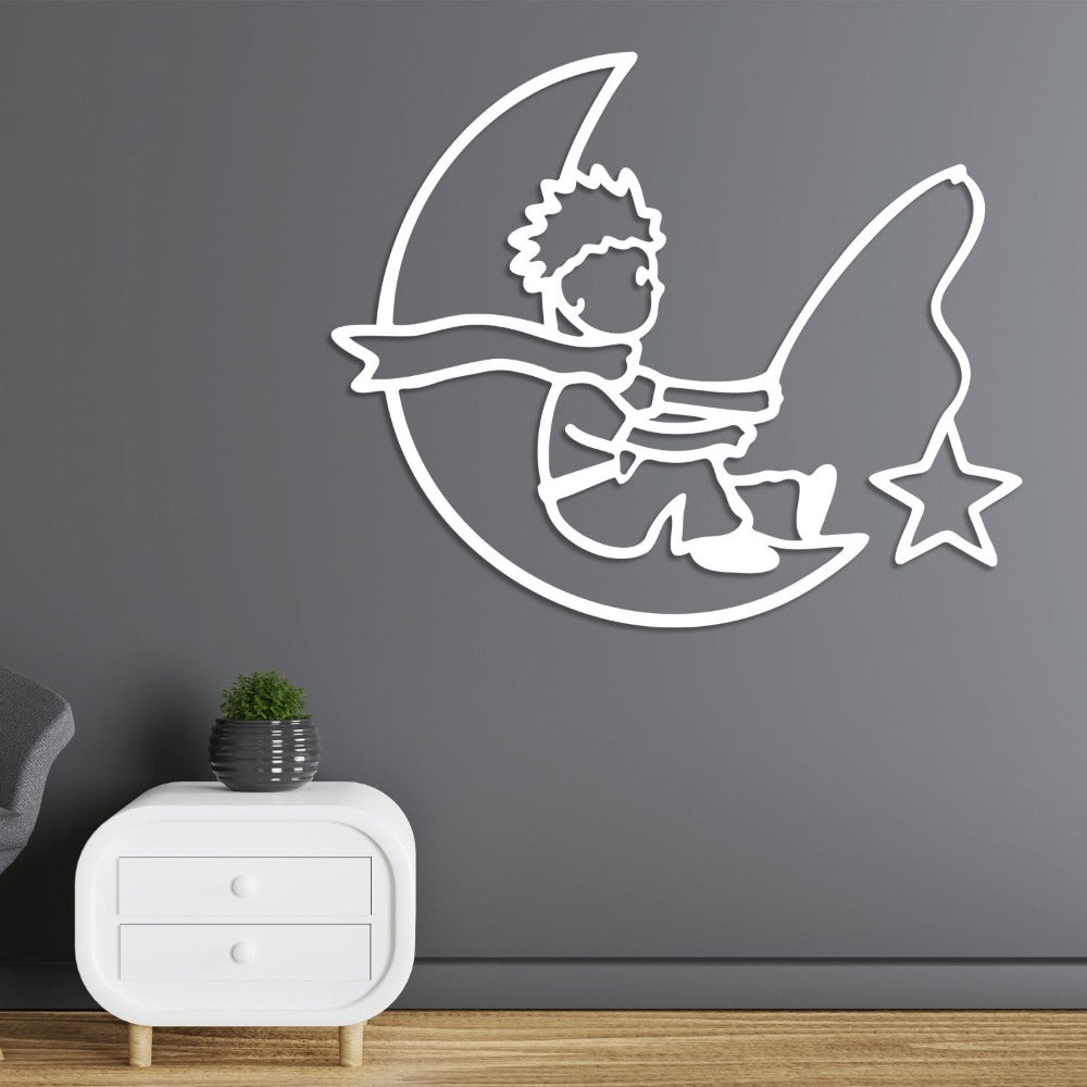 A white metal wall art of a little prince sitting on a crescent moon and fishing a star, mounted on a gray wall above a modern white nightstand.