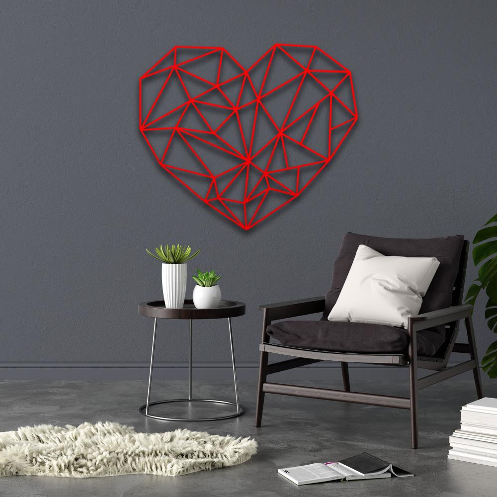 Modern geometric heart-shaped metal wall art in bold red color mounted on a gray wall above a cozy black chair with a white cushion, next to a round side table and decorative plants.