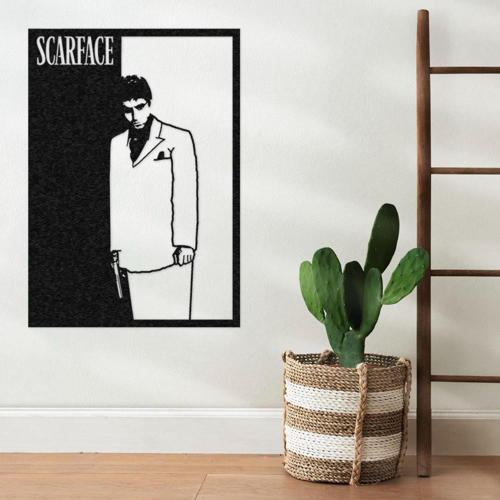 Scarface wall art showcasing a black and white silhouette of a man in a suit holding a gun, mounted on a light wall next to a decorative potted cactus in a woven basket and a minimalist wooden ladder.