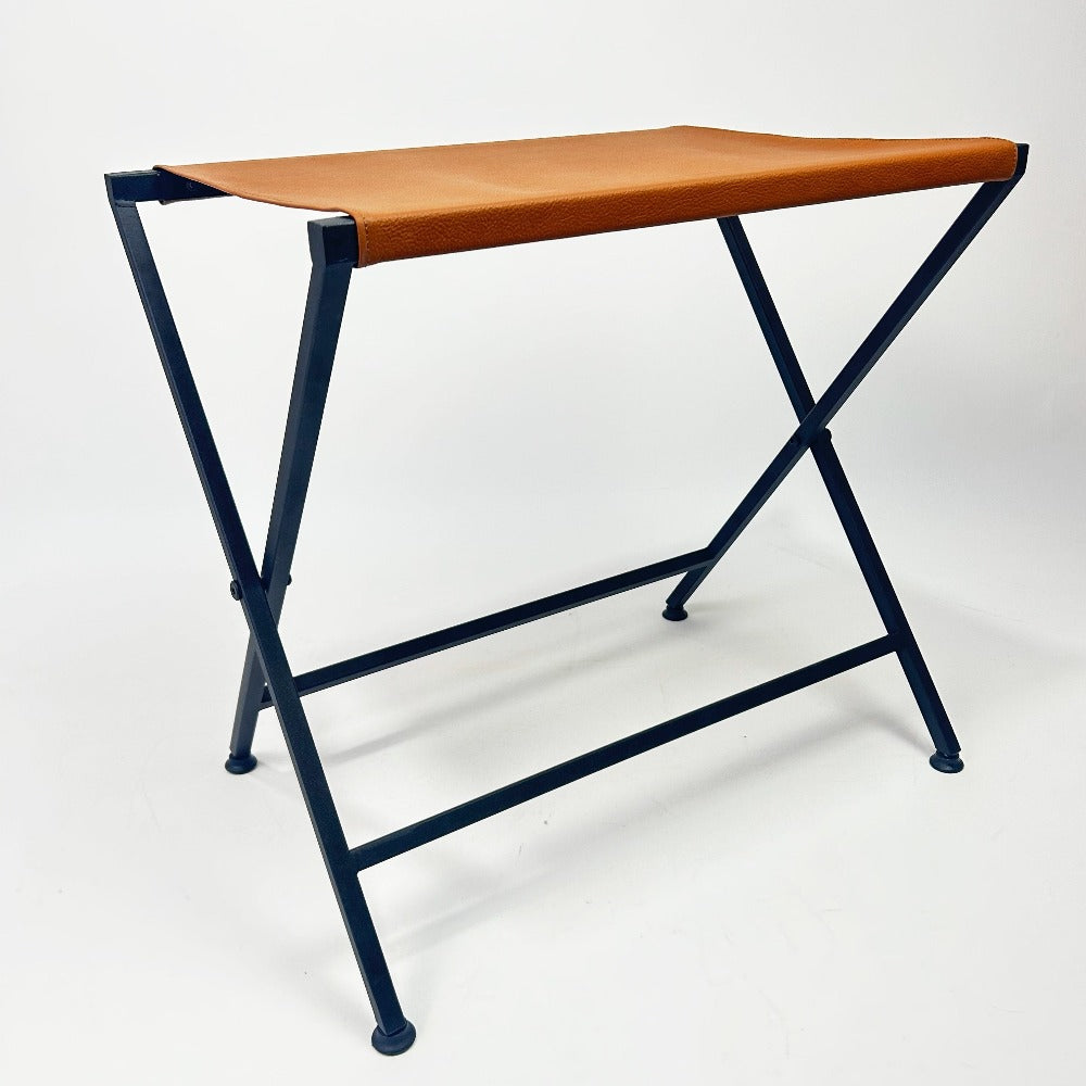 Side view of a foldable metal suitcase rack with a sturdy orange leather top, emphasizing its clean and modern aesthetics