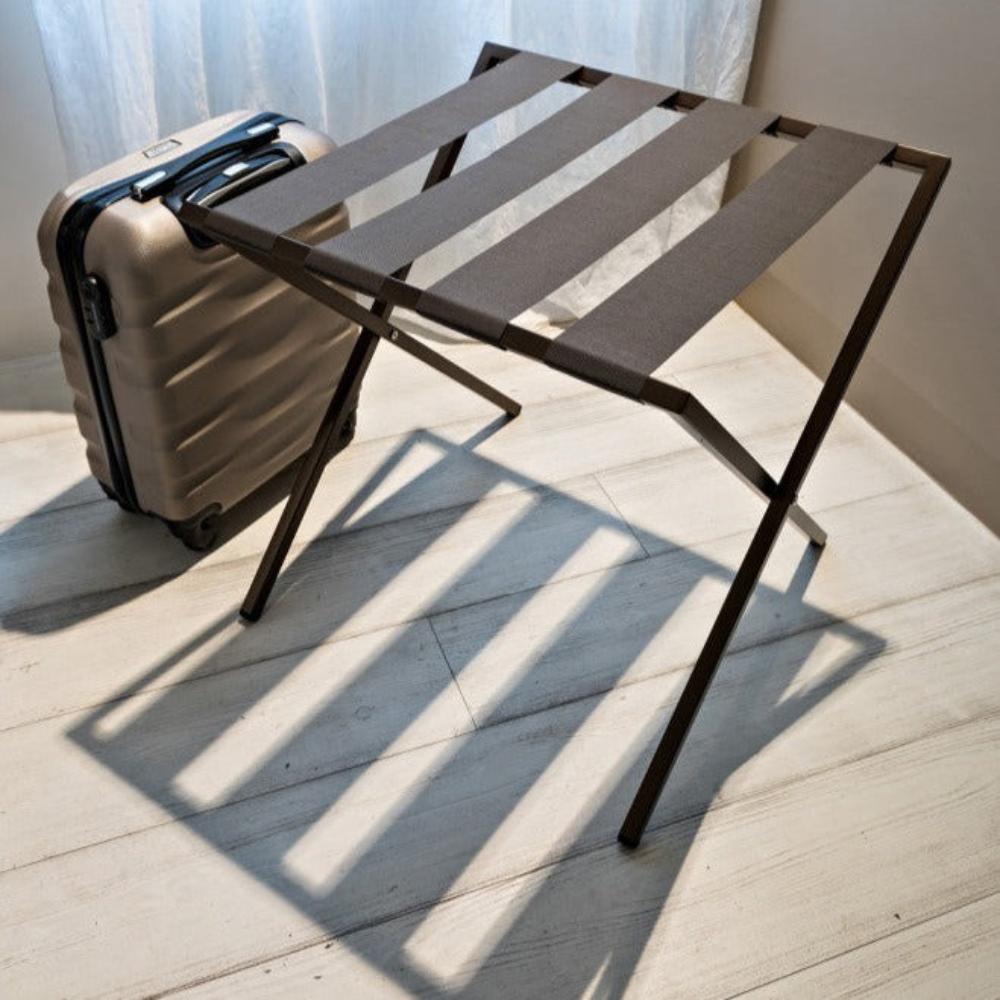 A bronze-tone foldable metal luggage rack with brown straps, paired with a beige suitcase in a softly lit room