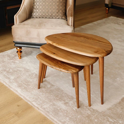 Three wooden nesting tables with a rounded design stacked together on a cream rug, placed near a beige armchair.