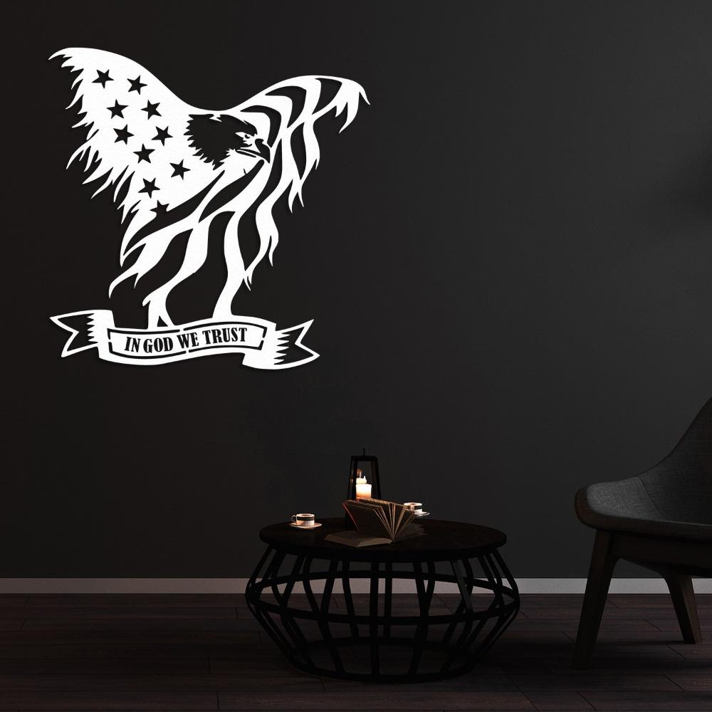White metal wall decor of a bald eagle with American flag-themed wings and the phrase "In God We Trust" on a ribbon, mounted on a dark wall above a modern black table with candles and books.
