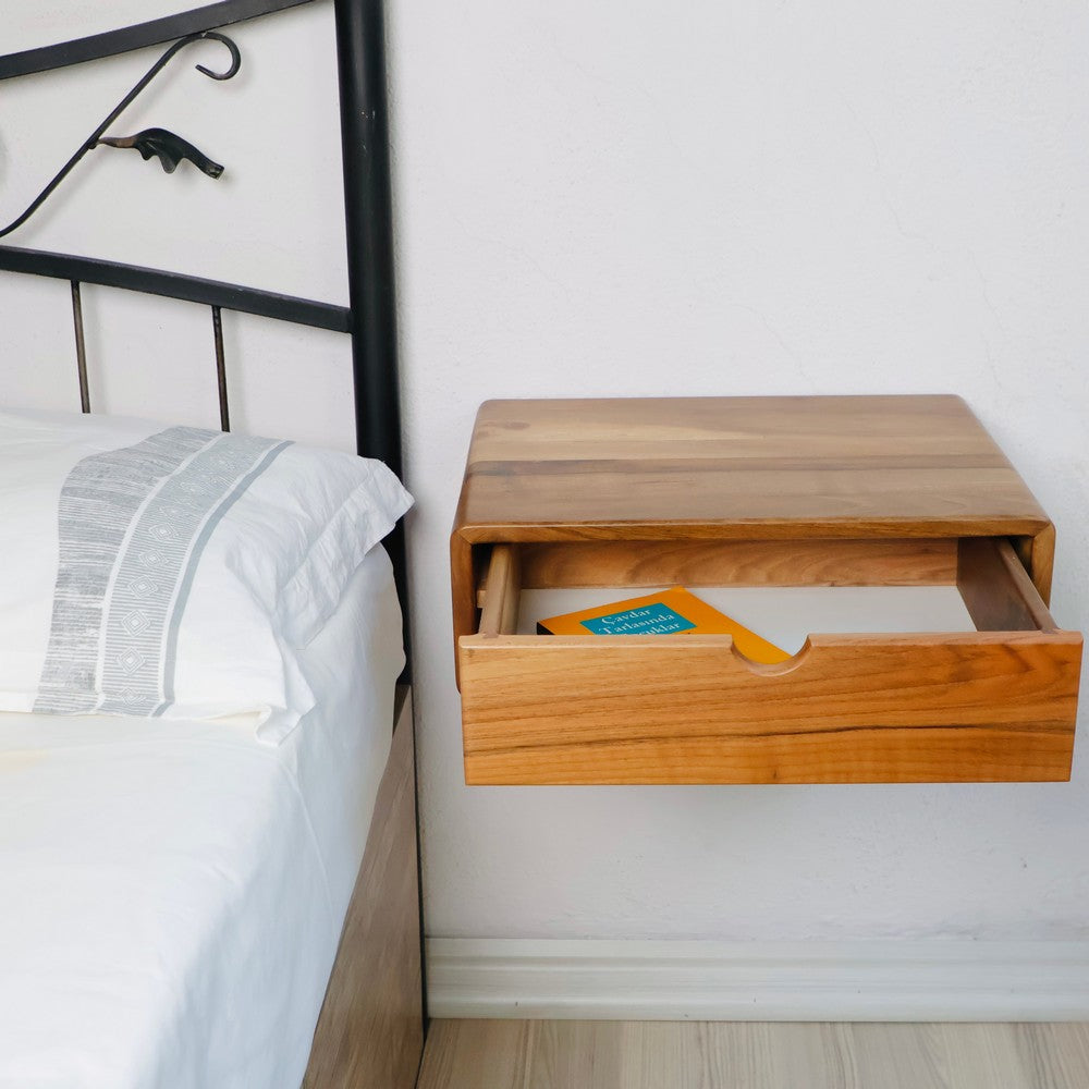 Wall-mounted wooden bedside table with its drawer partially open, revealing a book inside. The sleek natural wood finish complements the minimalist bedroom design.