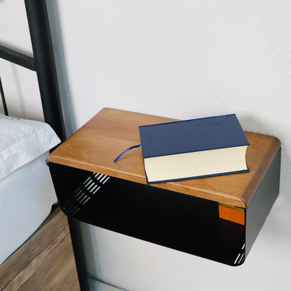 Close-up of a wall-mounted wooden and black metal bedside table. The table features a natural wood grain finish on top and a sleek black metal base with a storage compartment. A blue book is placed on the tabletop.