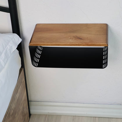 Wall-mounted wooden and black metal bedside table with a smooth natural wood top and a perforated black metal base. The minimalist design emphasizes functionality and modern aesthetics, shown for compact bedroom spaces.