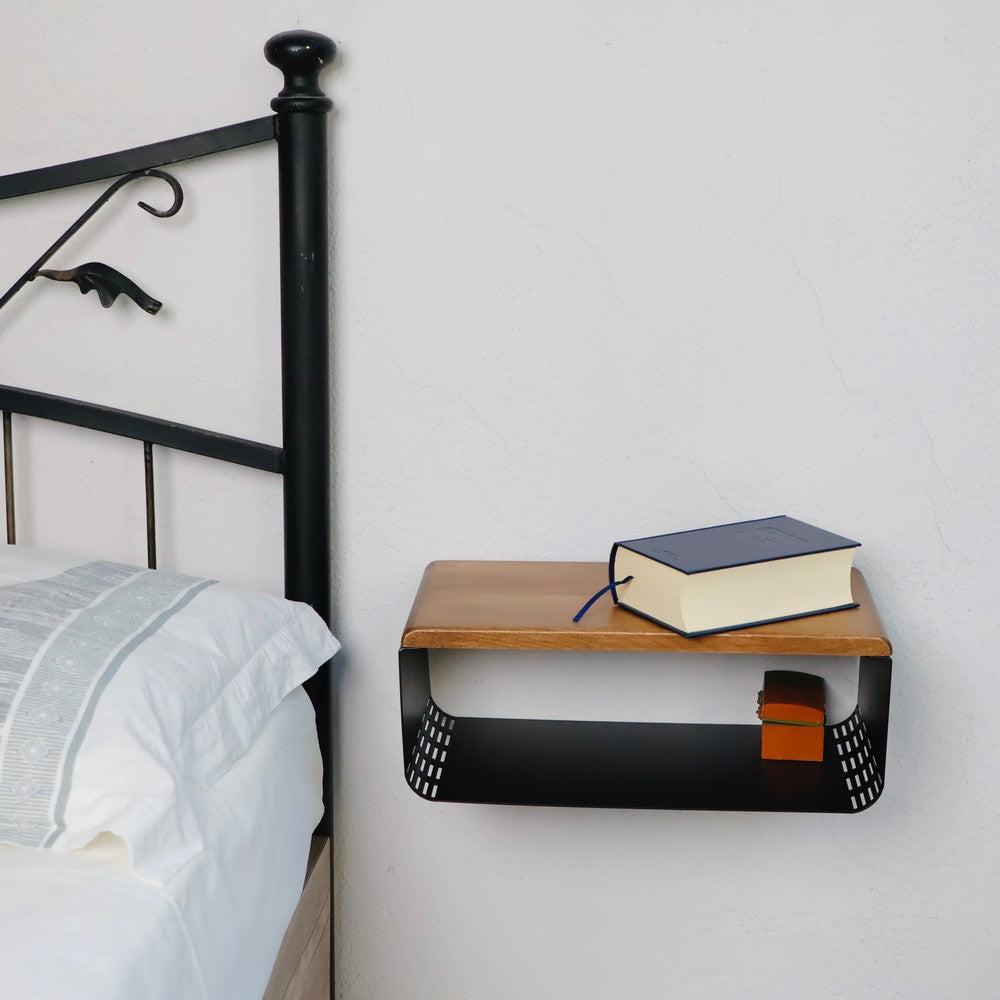 Wall-mounted wooden and black metal bedside table installed beside a bed. The table features a natural wood finish, a black metal base with a storage compartment, and a blue book on top, blending functionality with modern design.