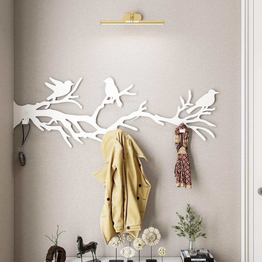 A white metal wall-mounted coat rack featuring birds perched on tree branches. The design showcases bird silhouettes sitting on elegant branches, providing both a functional hanging solution and a decorative touch. A beige coat and a patterned scarf hang from the hooks, while the scene is enhanced by minimalist decor elements below the rack.