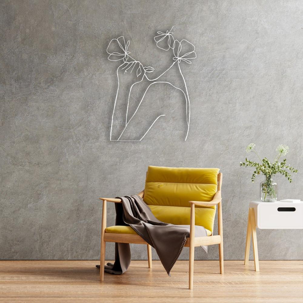 A white abstract metal wall art of a female figure with flowers displayed in a modern interior with a yellow accent chair and decorative items on a side table