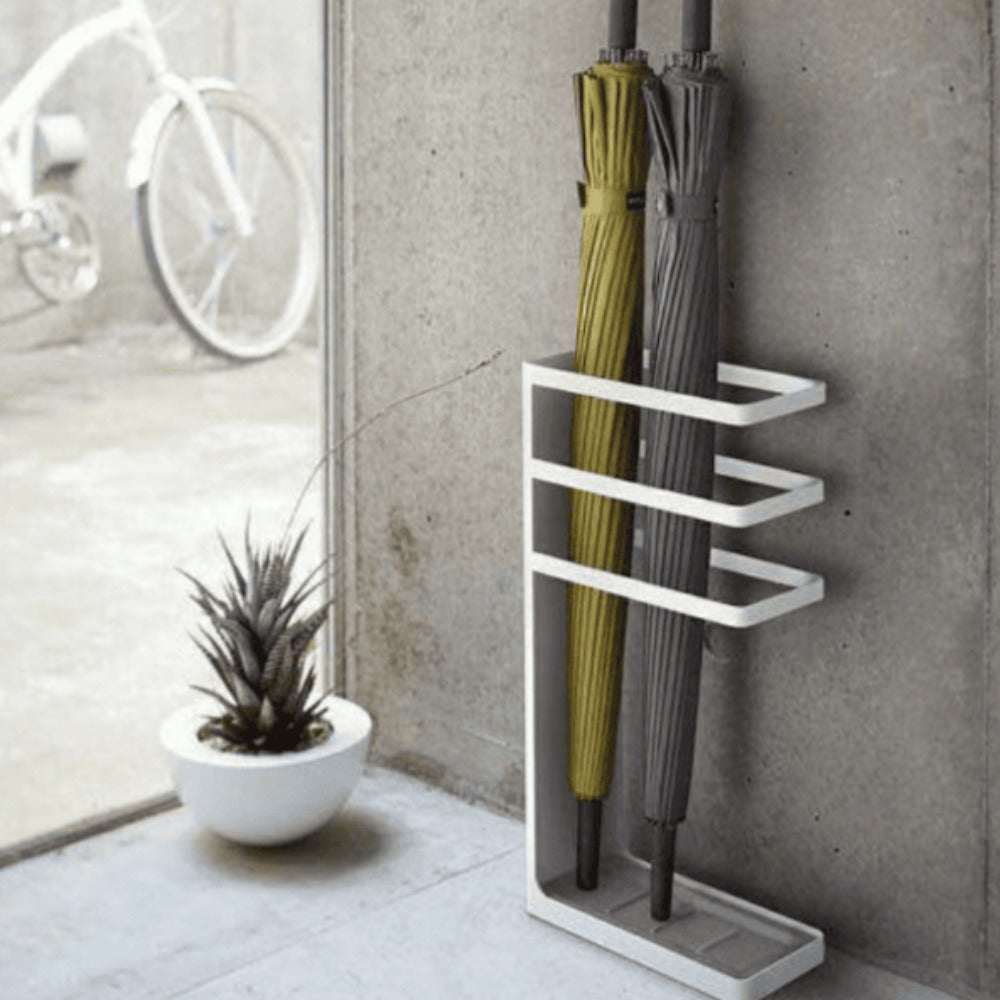 A white umbrella stand holding yellow and gray umbrellas, placed near a window with a small decorative plant and a concrete wall in the background