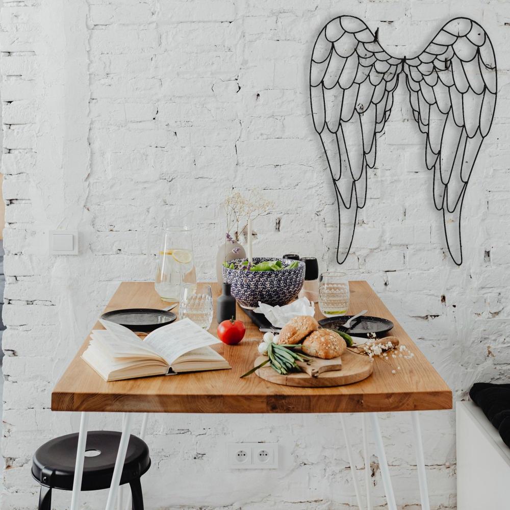 Metal angel wings wall art in black mounted on a soft beige wall, complementing a stylish modern interior with a tan leather chair, wooden console, and vibrant greenery.