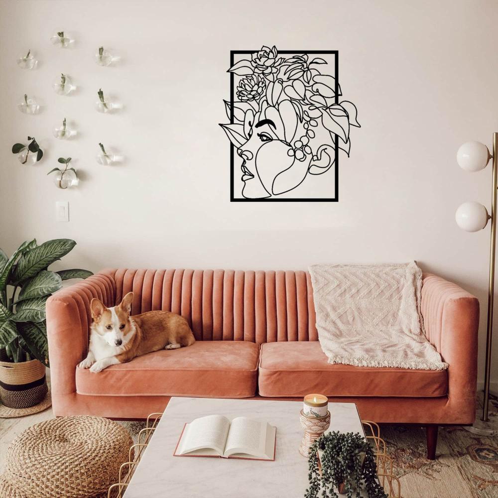 Metal wall art of a woman’s face with floral designs, displayed above a pink sofa in a bright and cozy living room with a corgi resting on the couch.