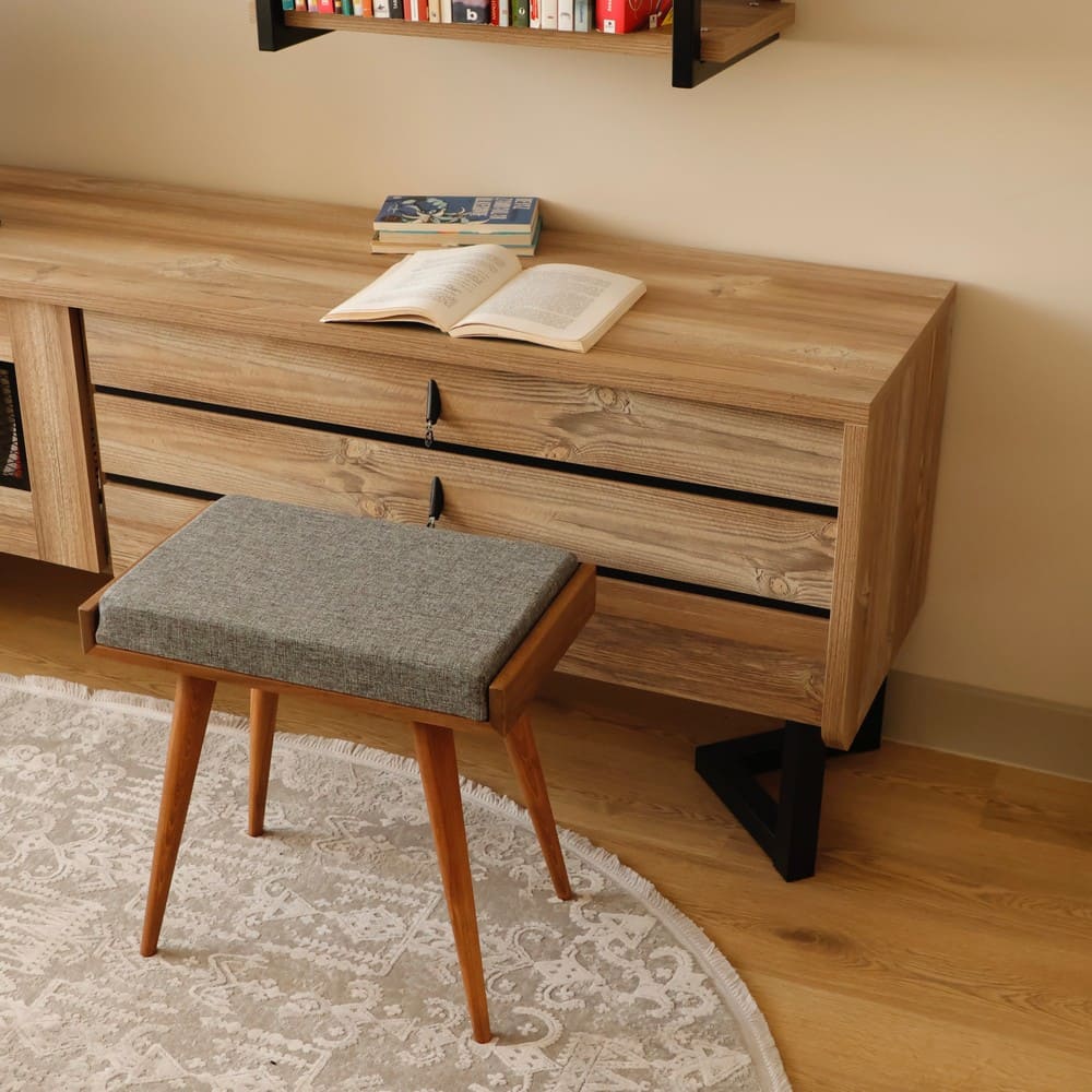 A compact wooden stool with a cushioned gray seat, placed in front of a modern wooden cabinet featuring open books and decorative items.
