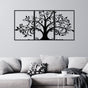 Three-piece black metal wall art featuring a tree with intricate swirling branches and leaves, mounted on a light gray wall above a modern light gray sofa. The sofa is styled with soft cushions, including one with abstract typography, and is accompanied by a small white round side table.