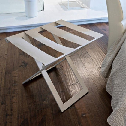 White metal foldable luggage rack with white genuine leather straps placed on dark wooden flooring next to a cream-colored chair, in a minimalist bedroom setup.