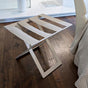 White metal foldable luggage rack with white genuine leather straps placed on dark wooden flooring next to a cream-colored chair, in a minimalist bedroom setup.