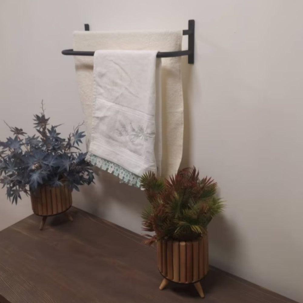 A decorative bathroom setup featuring a black towel holder with white embroidered towels and two potted plants in bamboo planters. The scene showcases a wooden countertop and a simple yet elegant arrangement for a cozy aesthetic.