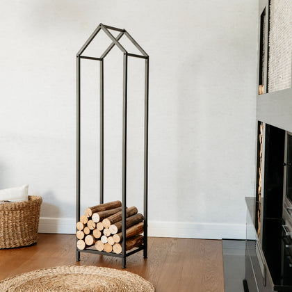 Black metal firewood rack with logs against a white wall in a room with a fireplace. house shaped firewood rack, decorative firewood rack for living room