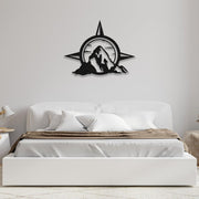 A compass and mountain design metal wall art mounted above a white bed with brown bedding in a simple, elegant bedroom.
