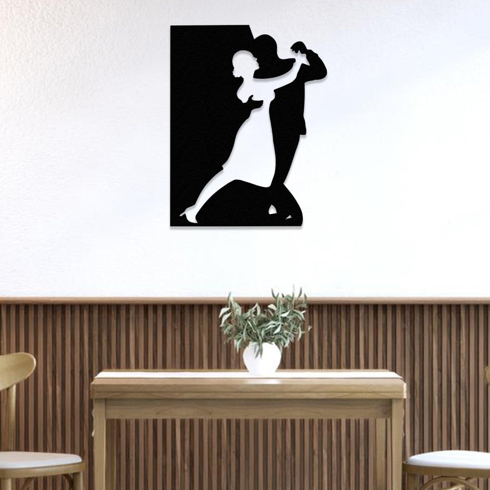A black metal wall art piece featuring a couple dancing, mounted on a plain white wall above a minimalist wooden table and a small plant in a white.