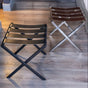 Two foldable luggage racks with brown genuine leather straps, one in matte black and the other in satin nickel finish, illuminated by warm ambient lighting on a wood-textured floor.