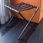 Black metal foldable luggage rack with brown genuine leather straps holding a beige suitcase in a softly lit guest-room interior.