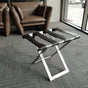 Foldable metal luggage rack with dark brown leather straps and a sleek metal frame, placed in a modern seating area with brown leather armchairs and gray carpet, offering a stylish and functional solution for luggage storage.