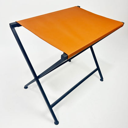 A foldable metal suitcase rack with a genuine leather top in orange, showcasing a sleek and metal design shown for guest rooms or travel use.