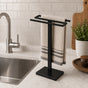 Black freestanding countertop double towel holder with hand towel beside a kitchen sink.