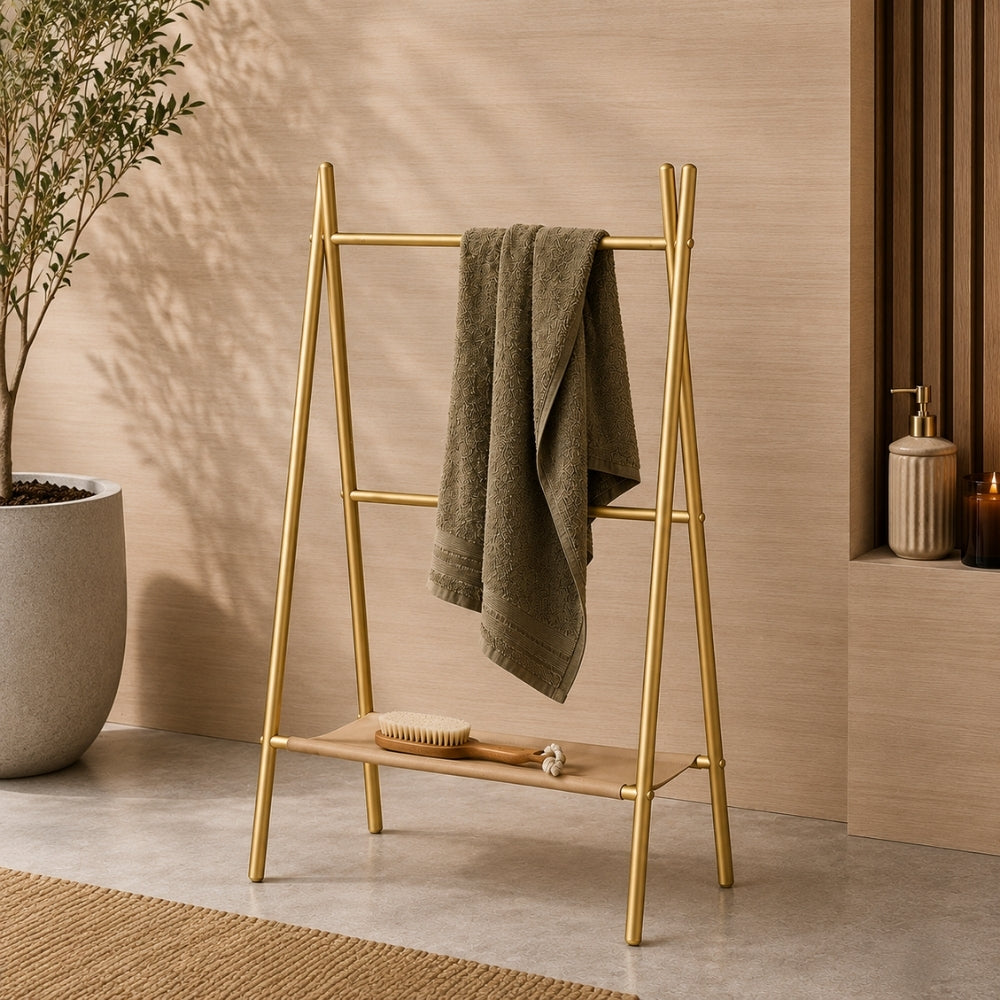 Gold freestanding ladder towel rack with shelf holding a green towel in a warm bathroom scene.