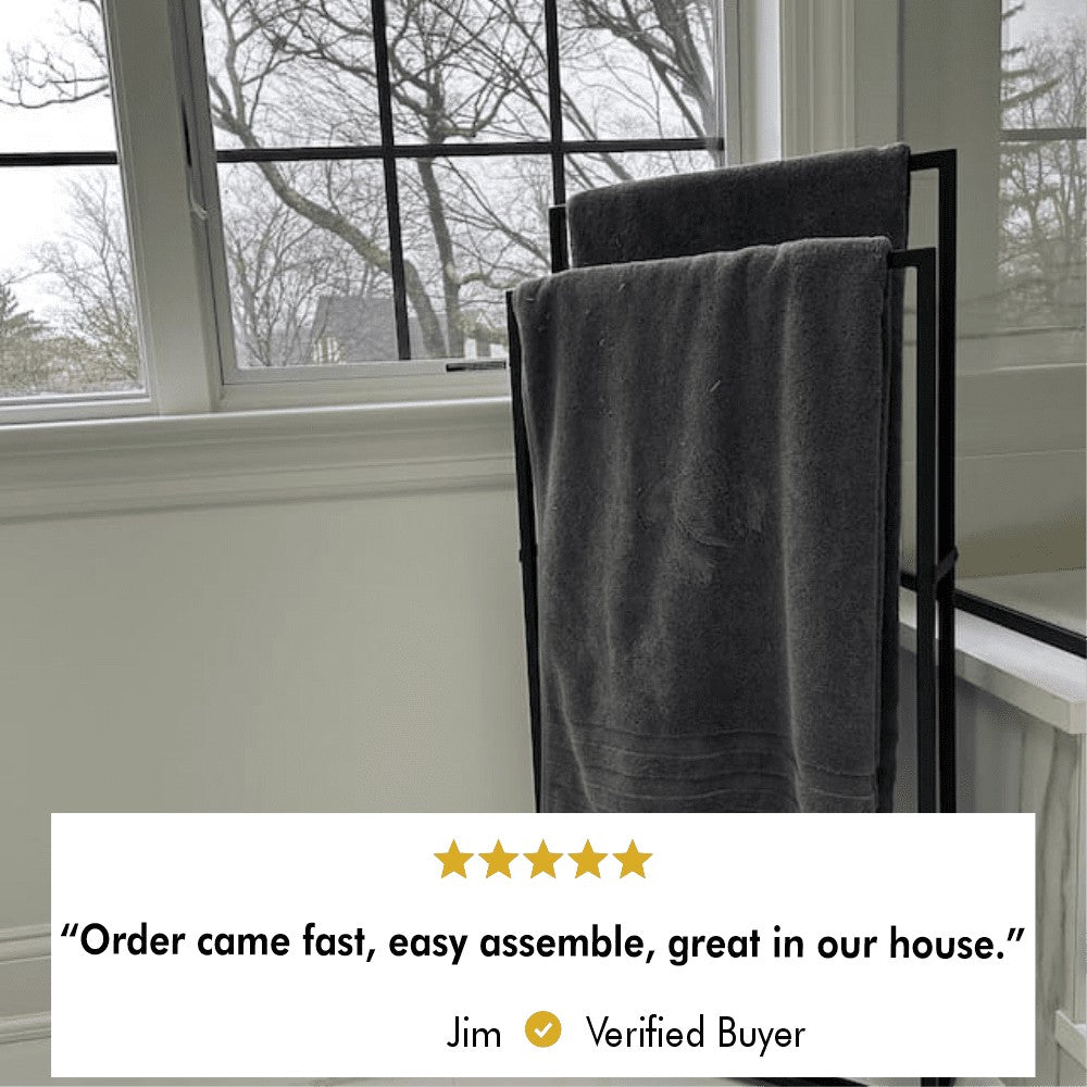 A black freestanding towel rack holding dark gray towels, placed in front of large windows with a view of bare winter trees.
