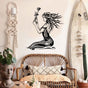 Elegant black metal wall art of a seated woman holding a rose, featuring intricate swirling patterns and flowing hair, displayed in a bohemian-style room. The decor includes a rattan bench with patterned cushions, a macrame wall hanging, a surfboard leaning against the wall, potted plants, and a decorative bust.