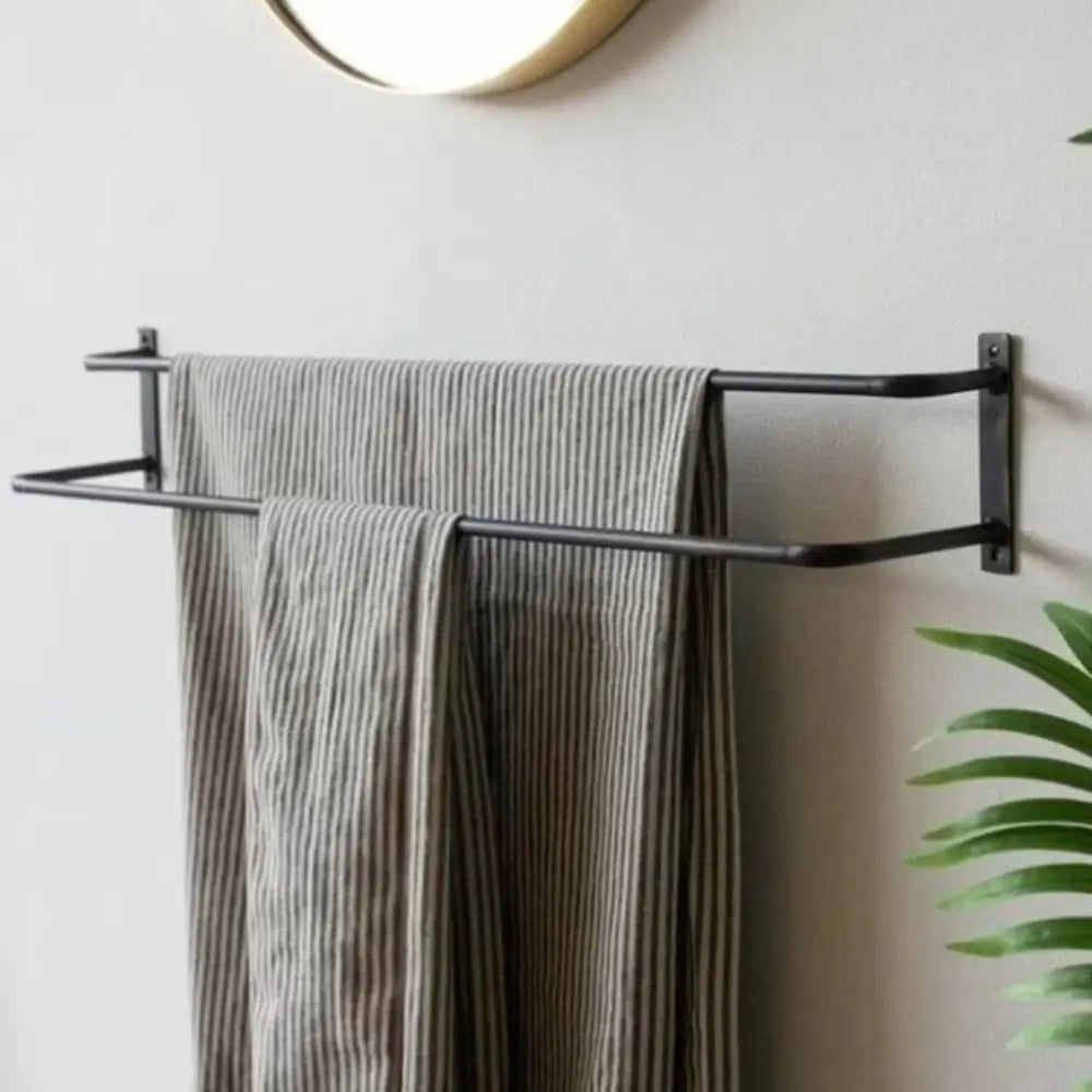 A close-up of a black towel holder mounted in a minimalistic bathroom, holding striped towels with a textured wall and a circular mirror in the background. Green plants add a refreshing touch to the clean and modern design.