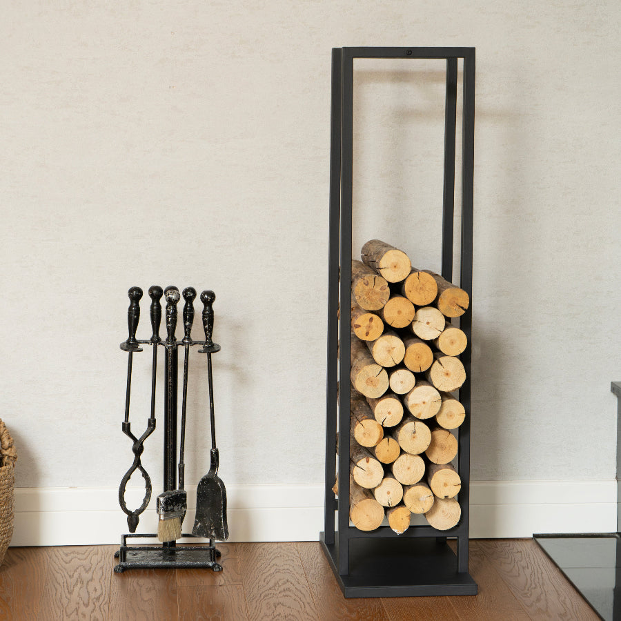 Black metal firewood rack with stacked logs and a set of black fireplace tools on a wooden floor.