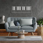 Piano keys-themed metal wall art in a sleek white design, mounted on a textured gray wall above a modern blue tufted sofa. The living space is complemented by wooden flooring, a round wooden coffee table with a flower vase, and minimalistic decor elements.